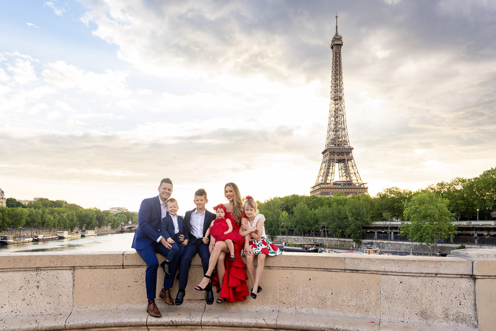 Elegant family of 6 wearing matchy red and blue outfits posing for family photos by the Eiffel Tower and seine river