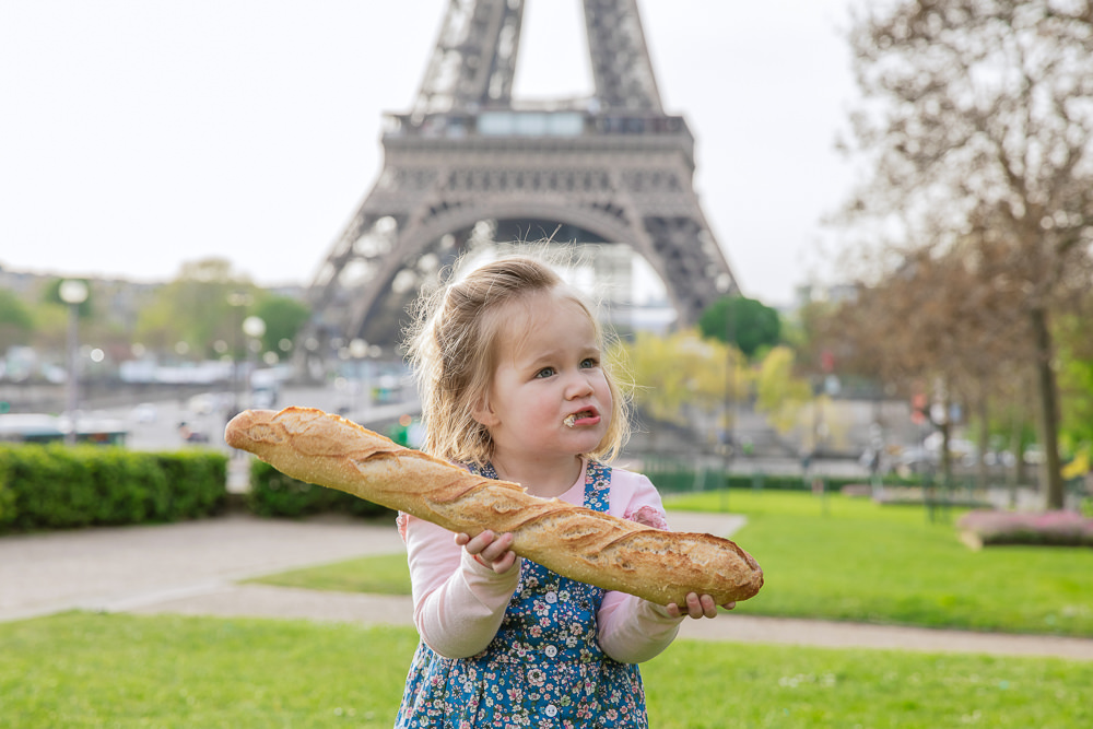 Little girl holding a baguette in her hands in front of the Eiffel Tower